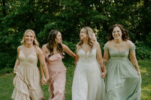 Bride and three bridesmaids in mismatched pastel dresses walk together smiling through green garden.