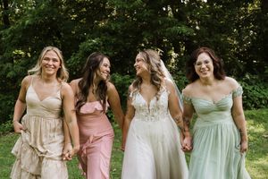 Bride in lace wedding gown walks with three bridesmaids in mismatched pastel dresses on green lawn.