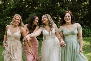 Bride and three bridesmaids in pastel dresses laughing and holding hands outdoors at a wedding.