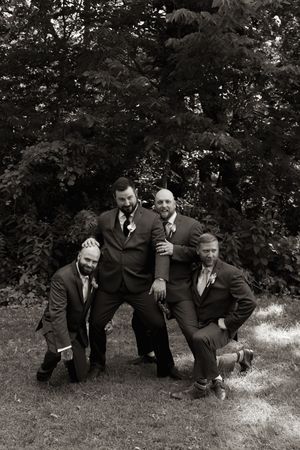 Four groomsmen in dark suits posing playfully in black and white photo outdoors near trees.
