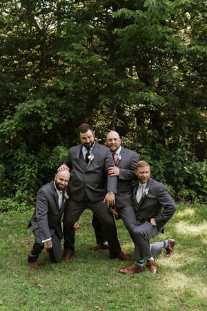 Four groomsmen in gray suits pose playfully together on green grass with lush trees in the background.