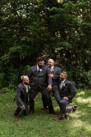 Groom and groomsmen in gray suits posing playfully on grass with lush green trees behind them.