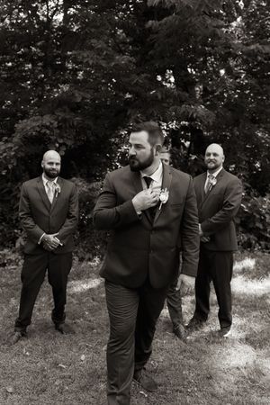 Groom adjusts boutonniere while groomsmen stand behind him outdoors in black and white wedding photo.