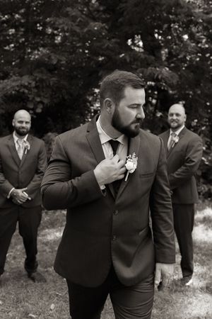Groom adjusting boutonniere in black and white photo with groomsmen standing behind him outdoors.