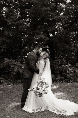 Black and white romantic photo of bride and groom kissing while holding floral bouquet in garden setting.