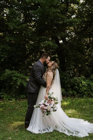 Bride and groom sharing a kiss outdoors, bride holding pink bouquet with long lace train.