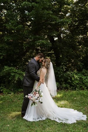 Bride and groom share romantic moment in garden, bride holding pink floral bouquet in elegant lace gown.