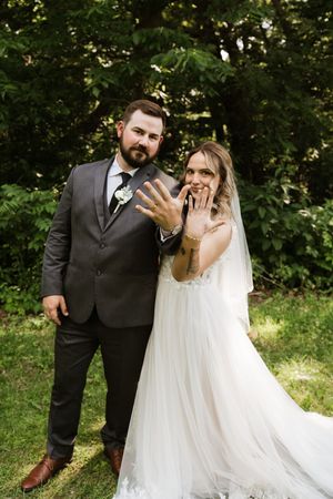 Smiling bride and bearded groom show off wedding rings outdoors in lush green setting.