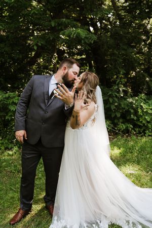 Bride and groom kiss while showing wedding rings in a lush green outdoor setting.
