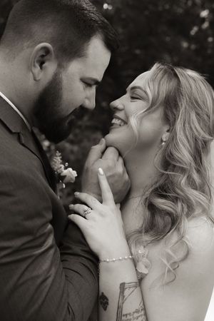 Black and white romantic moment of smiling bride and bearded groom touching foreheads closely.