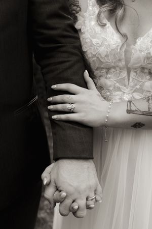Black and white close-up of bride and groom holding hands, showing wedding rings and tattoos.