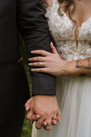 Close-up of bride and groom holding hands showing wedding rings, tattoos, and lace bridal gown details.