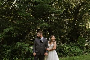 Bride in white tulle gown and groom in gray suit stand together among lush green trees.