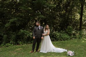Bride in white lace gown and groom in gray suit standing together in front of green trees.