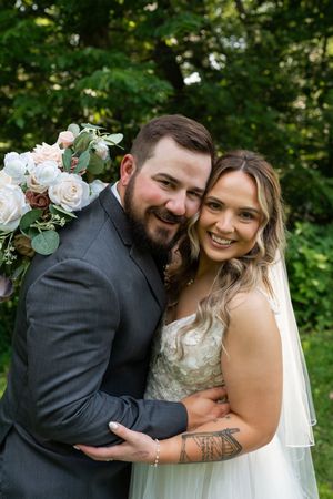 Smiling bride and groom pose together in front of lush green foliage with floral arrangement.