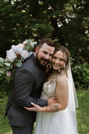 Smiling bride and groom embrace outdoors, bride holding white floral bouquet with eucalyptus, groom in gray suit.