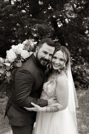Black and white portrait of smiling bride and groom embracing outdoors with floral bouquet.