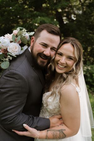 Laughing bride and groom embrace outdoors holding pink and white wedding bouquet.