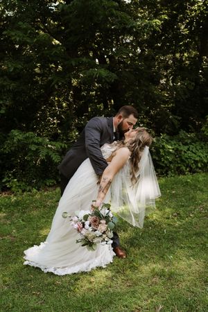 Groom dips bride for a kiss outdoors on grass, bride holding pink and white bouquet.