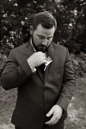 Bearded groom in dark suit smelling boutonniere in black and white wedding photo outdoors.