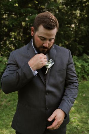 Bearded groom in gray suit adjusting white boutonniere lapel pin at outdoor wedding ceremony.