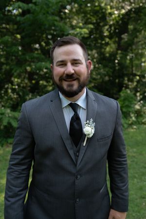 Smiling groom in gray suit with black tie and white rose boutonniere posing outdoors among green trees.