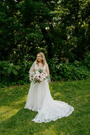 Bride in lace wedding gown holding pink bouquet standing on green lawn surrounded by lush trees.