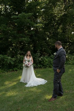 Groom in gray suit watches smiling bride in white lace gown holding pink and white bouquet during outdoor first look.