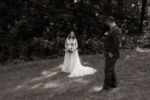Black and white first look photo of bride in flowing gown holding bouquet facing groom outdoors.