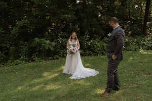 Bride in white gown with train holds bouquet during first look with groom on green lawn.
