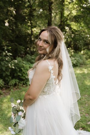 Bride in lace wedding gown and veil smiles over shoulder in lush garden setting.