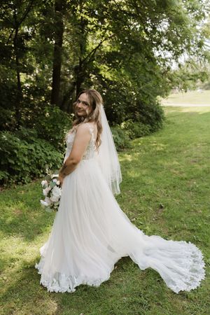 Bride in elegant white lace tulle gown with veil and train poses outdoors on green lawn holding bouquet.