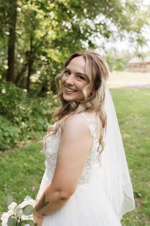 Smiling bride in white lace gown and veil holds bouquet in sunlit garden.