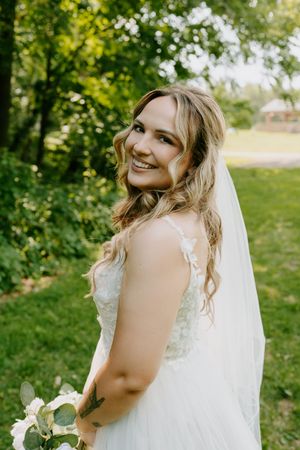 Smiling bride in lace wedding gown with veil and tattoo holding bouquet in lush green garden.