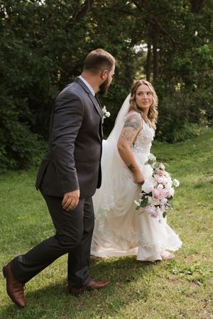 Bride and groom walk hand in hand on grass, bride glancing back holding floral bouquet.