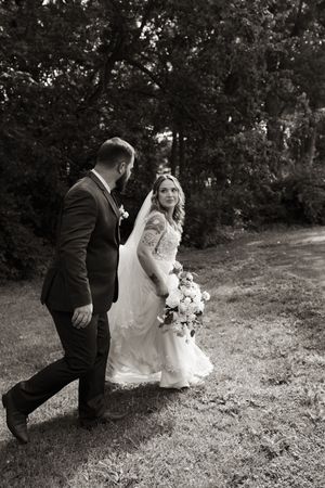 Black and white photo of bride in lace gown and groom walking together on grassy lawn.