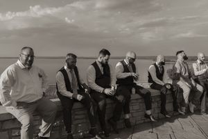Black and white photo of groomsmen relaxing on stone wall outdoors before wedding ceremony.