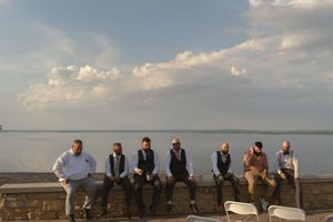 Seven groomsmen in suits sit on stone wall with scenic lake and cloudy sky backdrop at outdoor wedding.