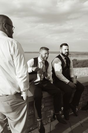 Black and white photo of groomsmen in vests relaxing on a stone wall with a scenic water view.