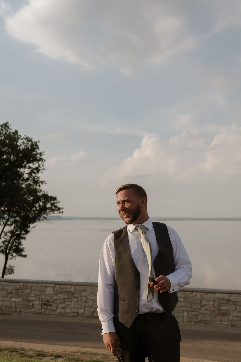 Handsome bearded groom in vest and tie smiles outdoors by lakeside on wedding day.