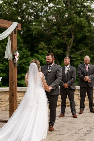 Bride in a flowing white veil holds hands with a bearded groom in gray suit at an outdoor wedding ceremony.