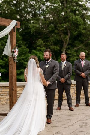 Bride and bearded groom hold hands at an outdoor wedding ceremony with groomsmen standing nearby.