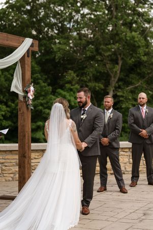 Bride and groom holding hands at outdoor wedding altar with groomsmen standing behind under wooden arch.