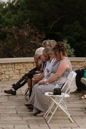 Wedding guests seated on white folding chairs at outdoor stone patio ceremony, emotional family members embracing.