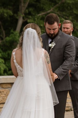 Bride and groom hold hands during outdoor wedding ceremony, bride in lace veil and gown, groom in gray suit.