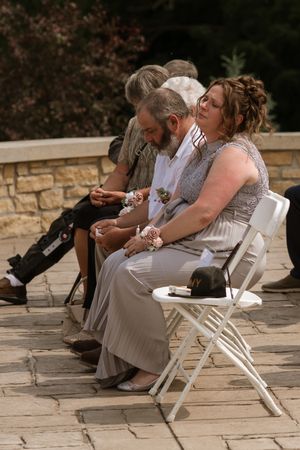 Wedding guests in formal attire seated on white chairs during outdoor ceremony on stone patio.