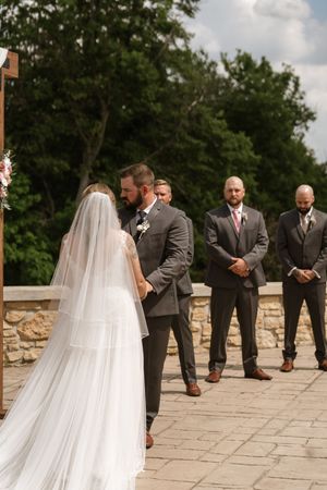 Bride and groom exchange vows at an outdoor stone patio wedding with groomsmen standing in the background.