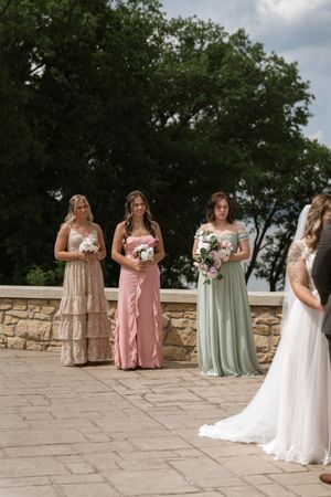 Three bridesmaids in mismatched pink and green gowns holding bouquets stand beside a bride at an outdoor wedding.