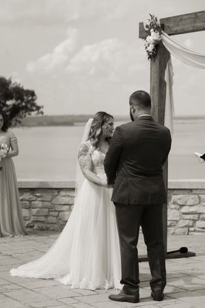 Black and white photo of bride and groom exchanging vows at outdoor waterfront wedding ceremony under wooden arch.