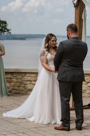Bride with tattoos in lace wedding gown holds grooms hands during outdoor lakeside wedding ceremony.
